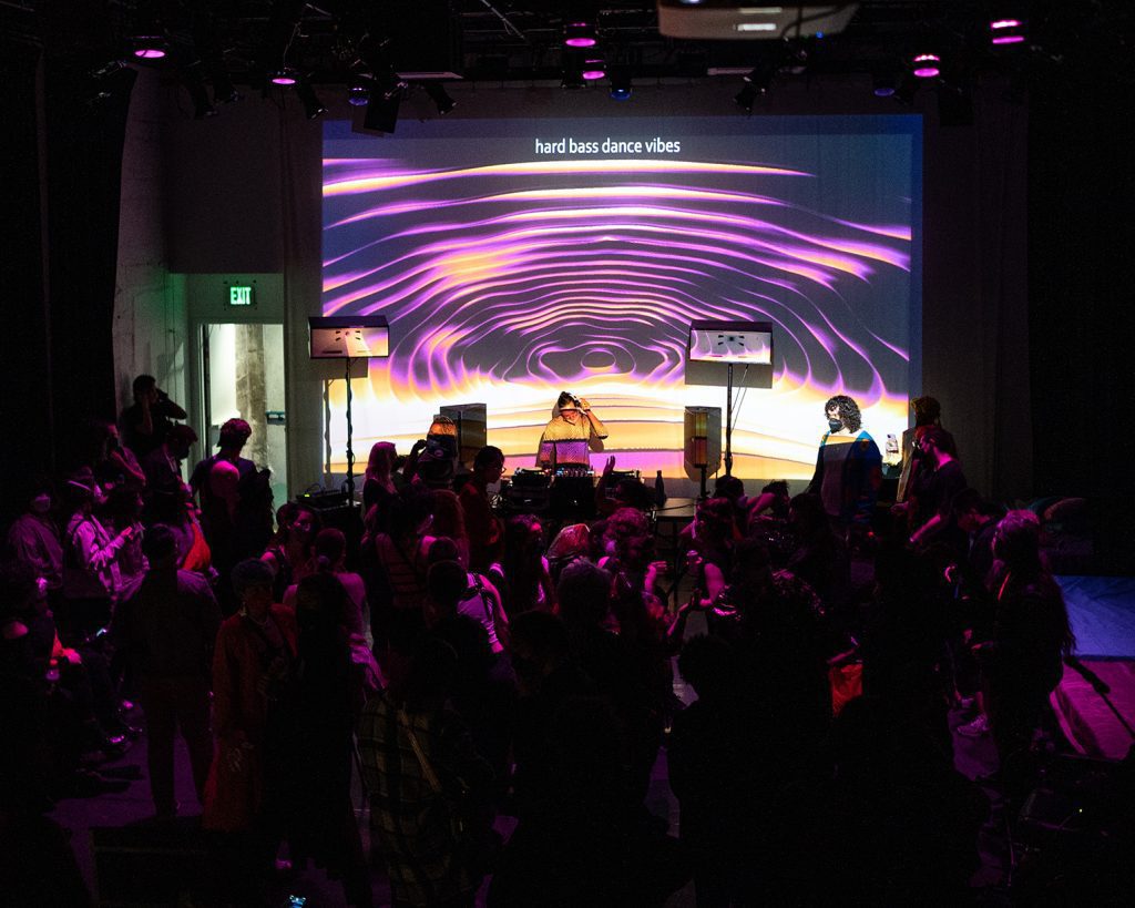 A crowd of disabled and non-disabled partygoers dance at a nightlife event. Behind a DJ booth, purple abstract illustrations are placed beside text that reads "Hard bass dance vibes"