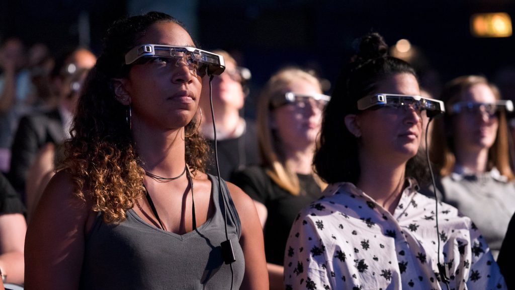 A group of diverse theater patrons watches a play with high tech captioning glasses that allow them to see both captions and the theater in their frame of vision.