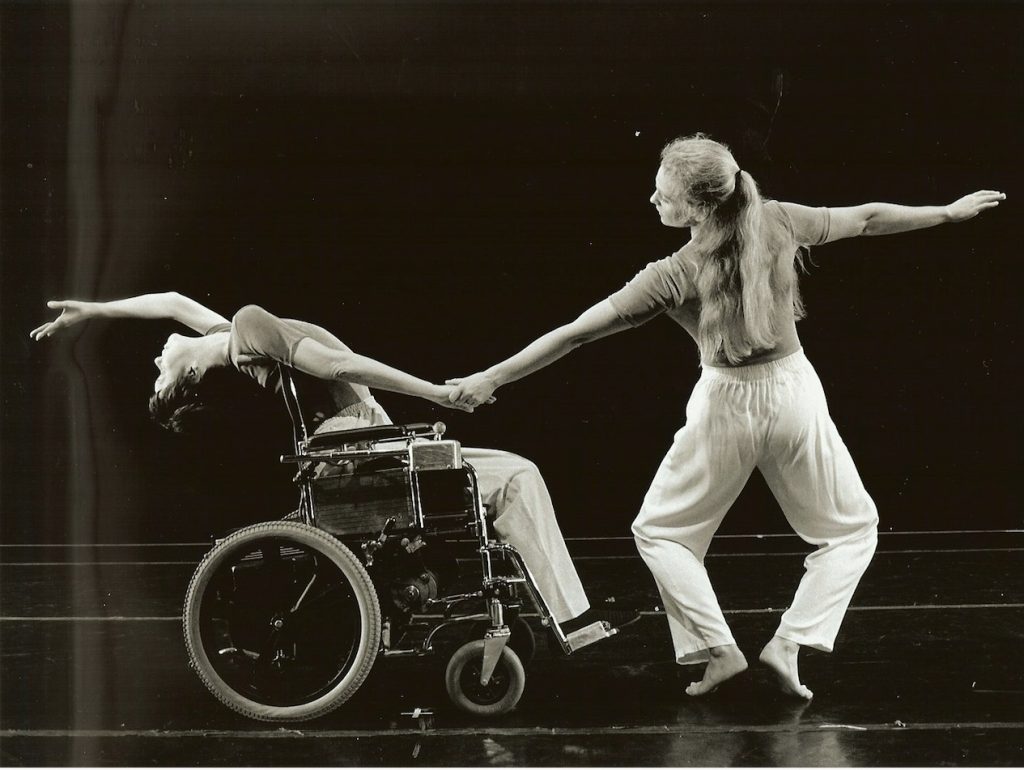 A black and white photo of Megan and Stephanie. Megan leans back in her wheelchair with her arm outstretched. Stephanie holds Megan’s other arm while she pliés