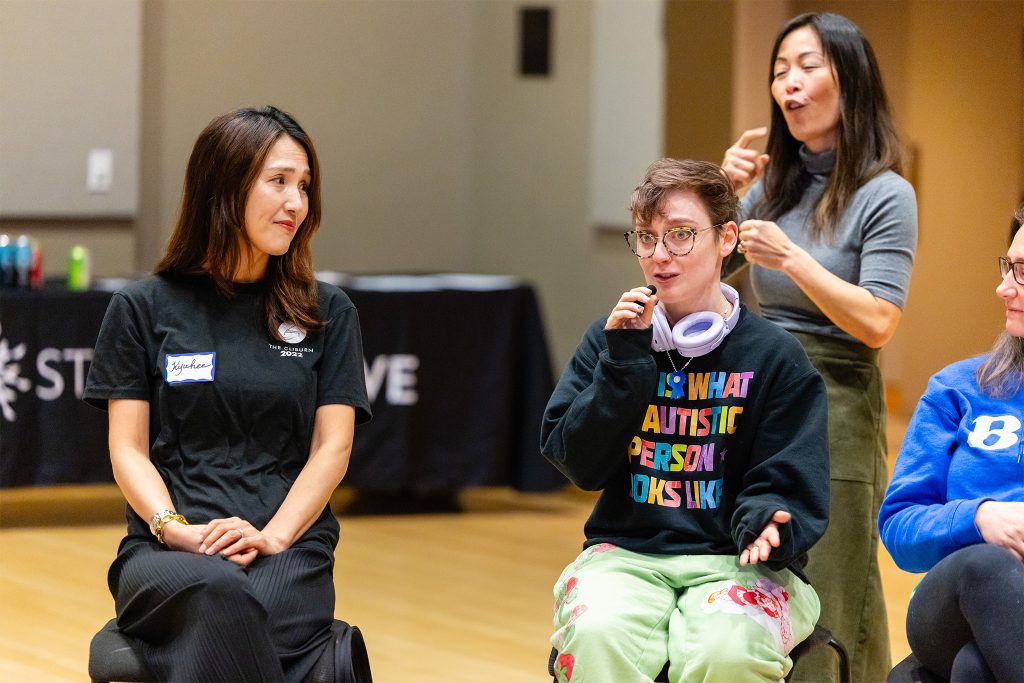 An AXIS teacher training participant speaks into a microphone, while another participant listens with compassion. An ASL interpreter signs behind them. The speaker wears a shirt that says "This is what an autistic person looks like."