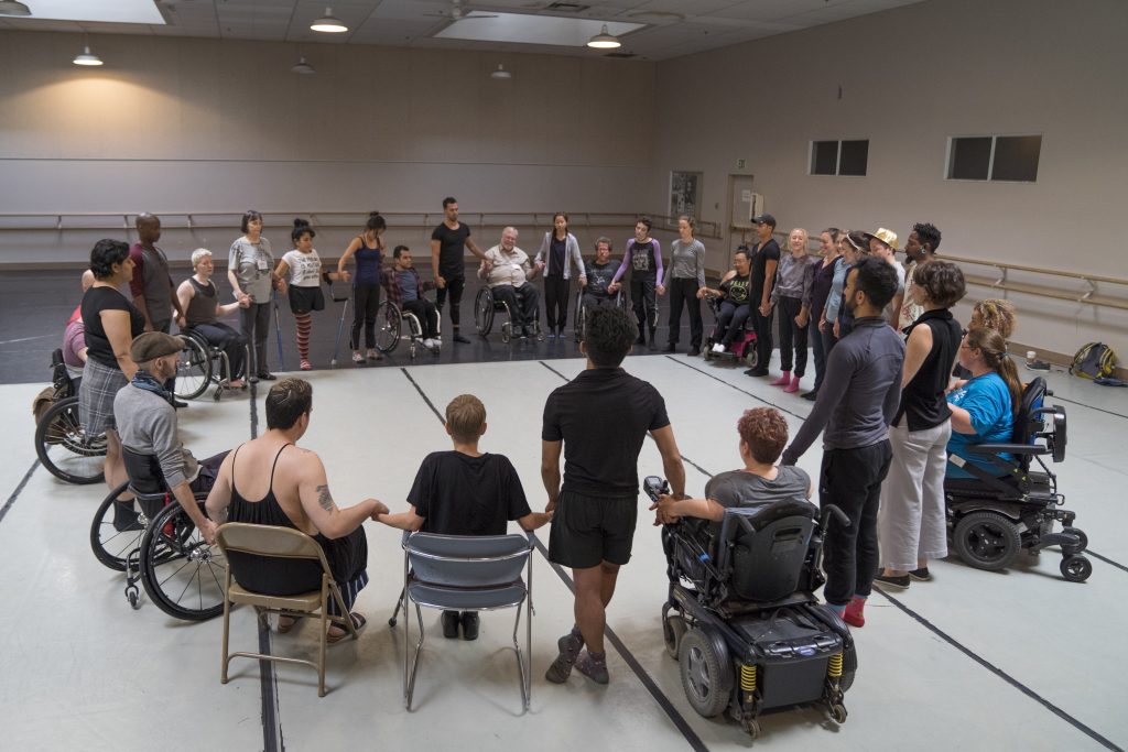 A diverse group of disabled and non-disabled individuals all hold hands in a large circle. Some people sit in chairs or their wheelchairs; others are standing.