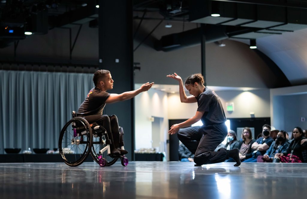 On a dance stage, JanpiStar reaches forward in their wheelchair with one arm while Alaja Badalich crouches down and lifts one arm up towards Janpi.