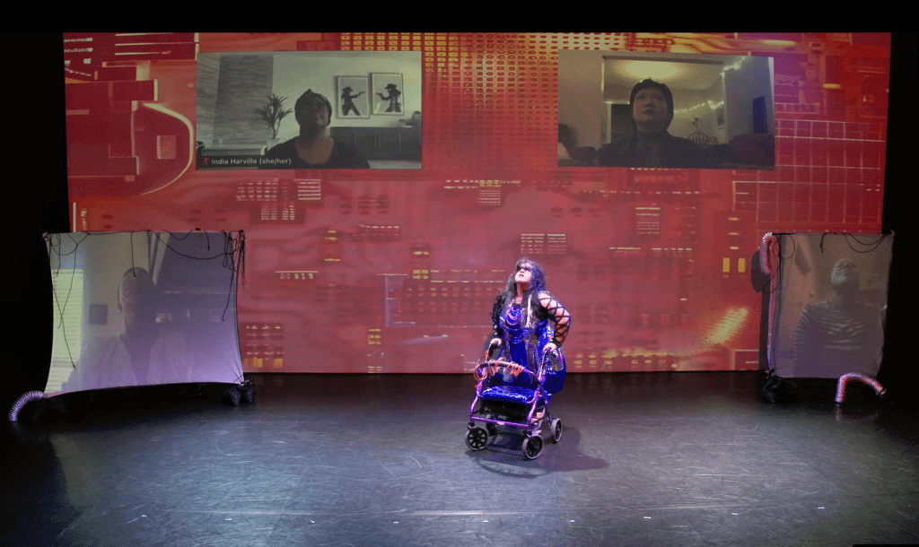 A photo of Vanessa who is a Chicana woman who has caramel skin is standing on stage looking up. She is wearing a blue and silver holographic dress with tubes on it. Her purple walk is in front of her. There are four disabled dancers in four different projections screen dancing with her, looking up. The projection background has images of messy wires & computer hardware. From left to right: Em Waters who is a light skinned Black person with short black Afro, wearing a pink face mask and a white top, India Harville who is a Black, queer, Disabled cis woman wearing a black head wrap and black top, 陳璐/Lu Chen who is a light-skinned East Asian person with short black hair wearing a blazer, & Ande Diedjomahor who is a Black person with a striped black and white shirt.