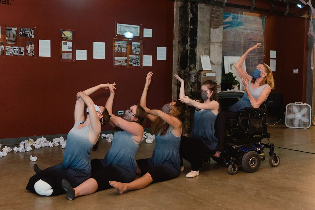 Five disabled and non-disabled dancers sit on the floor and their wheechairs in a line, rising upper limbs with one hand on elbows in energetic unison. They wear blue-white gradient tops and face masks.