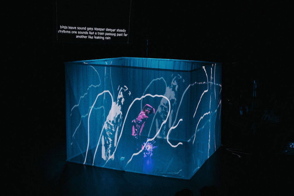 YoYoPhoto_Cropped In a blackbox theater, a fabric cube is luminescent in blue and purple with white gesture drawings from Pelenakeke Brown, drawing with live projections. Inside the cube, Yo-Yo arches to the side, ther arm over her head, shaping her body to the looping drawings on the screen. Behind her a description of the sound “birds leave sound gets deeper deeper steady rhythms are sounds like a train passing past far another like leaking rain.”